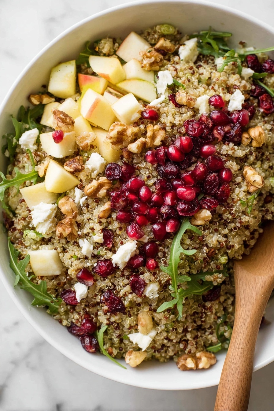 A large white bowl filled with a colorful quinoa salad sits on a white marbled surface. The salad has several layers visible on top: the main layer is light brown quinoa mixed evenly with scattered bright red pomegranate seeds, small chunks of pale yellow apple, white crumbled cheese, dark red dried cranberries, and green arugula leaves spread across. Some walnut pieces are also mixed in, adding texture. A wooden spoon with a smooth, light brown finish is partially inside the bowl, resting on the right side, slightly lifting some of the salad. Nearby, a few arugula leaves and quinoa grains are scattered on the white marbled surface along with a halved pomegranate showing its red seeds inside. Photo taken with an iphone --ar 2:3 --v 7