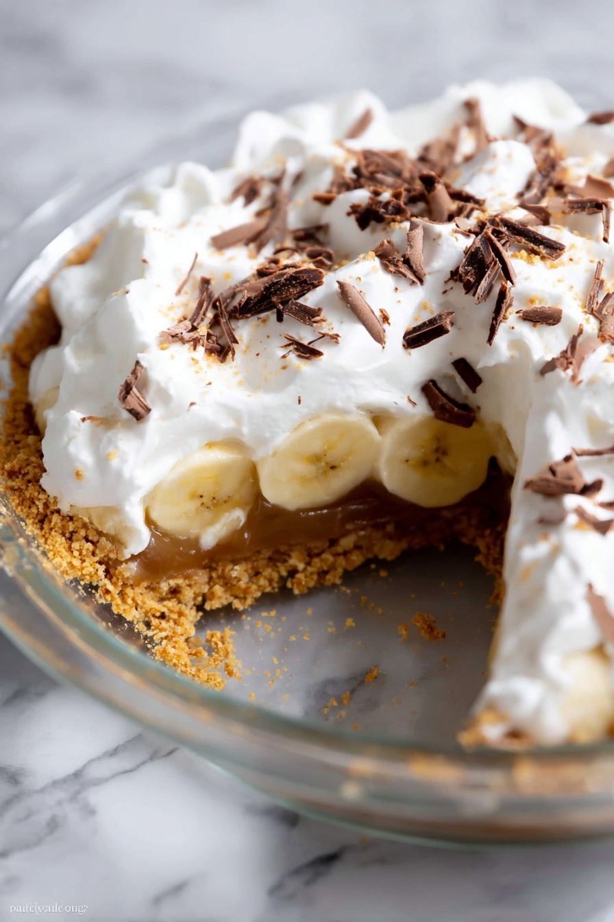 This image shows a close-up of a three-layer pie in a clear pie dish on a white marbled surface. The bottom layer is a golden brown crumbly crust, the middle layer has light yellow banana slices arranged in a single layer above a smooth caramel filling, and the top layer is thick, fluffy white whipped cream with dark brown chocolate shavings scattered on top. A wedge of the pie is missing, revealing the layers clearly. The texture of the whipped cream looks soft and slightly peaked, and the bananas look ripe but firm. photo taken with an iphone --ar 2:3 --v 7