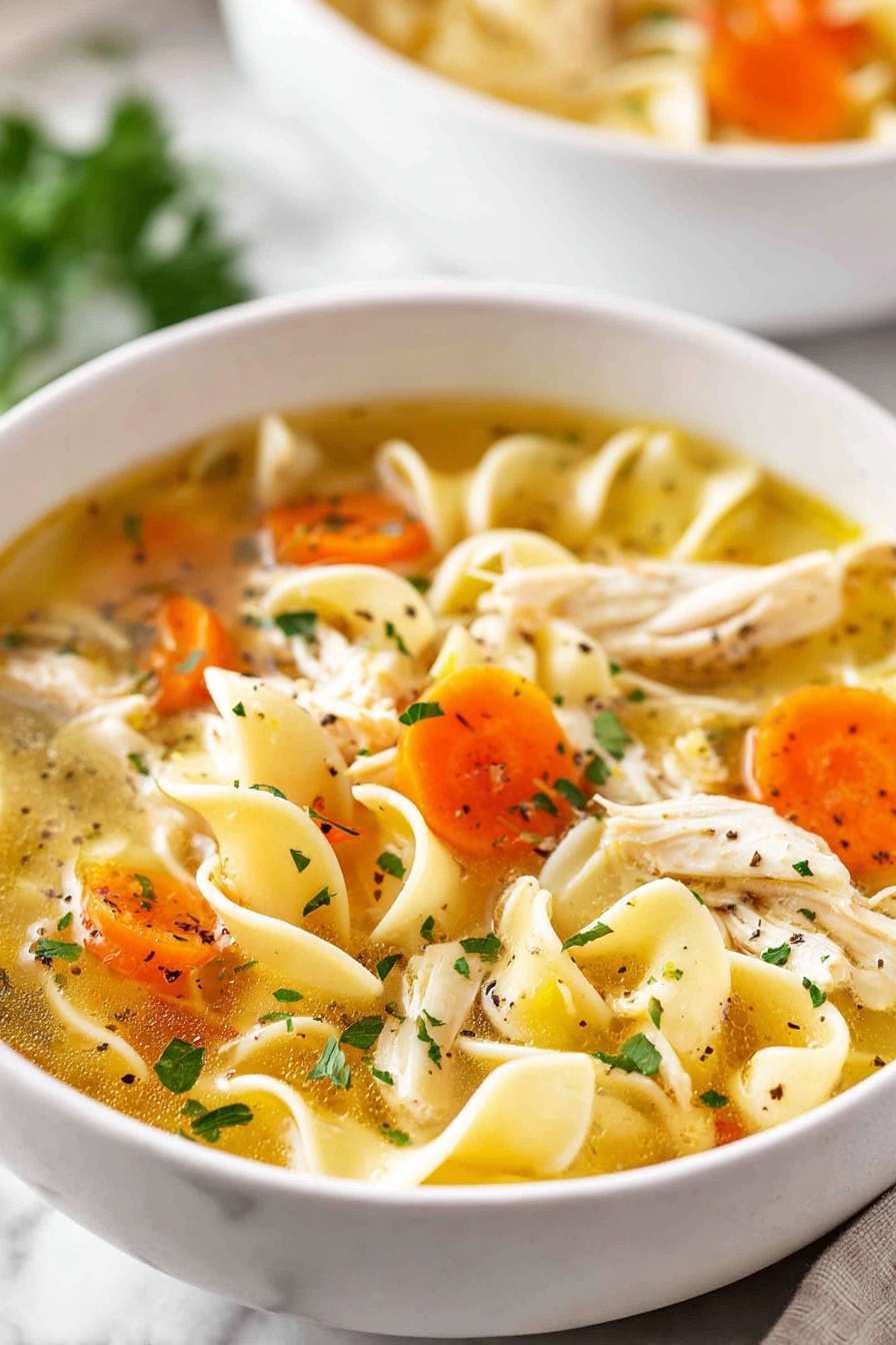 A white bowl full of chicken noodle soup held by a woman's hands with glittery pink nail polish, showing three main layers: a clear golden broth with small herbs and black pepper, bright orange carrot slices and light green celery pieces floating in the soup, and shredded white chicken mixed with wide, wavy egg noodles filling the bowl. The soup looks warm and fresh, with a white marbled surface visible around the bowl and a silver spoon beside it. photo taken with an iphone --ar 2:3 --v 7