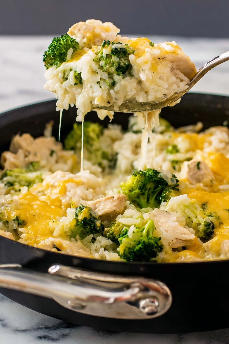 This image shows a creamy casserole in a round pan with layers of soft white rice mixed with light beige chicken pieces and bright green broccoli florets. The top layer is a melted yellow and white cheese blend that covers the rice and broccoli, with some fresh green parsley sprinkled on top. A shiny silver spoon is scooping a portion, showing the same layers close together inside it. The pan sits on a dark textured surface that is changed to white marbled texture. The food looks warm and cheesy with a mix of smooth and slightly textured parts. photo taken with an iphone --ar 2:3 --v 7