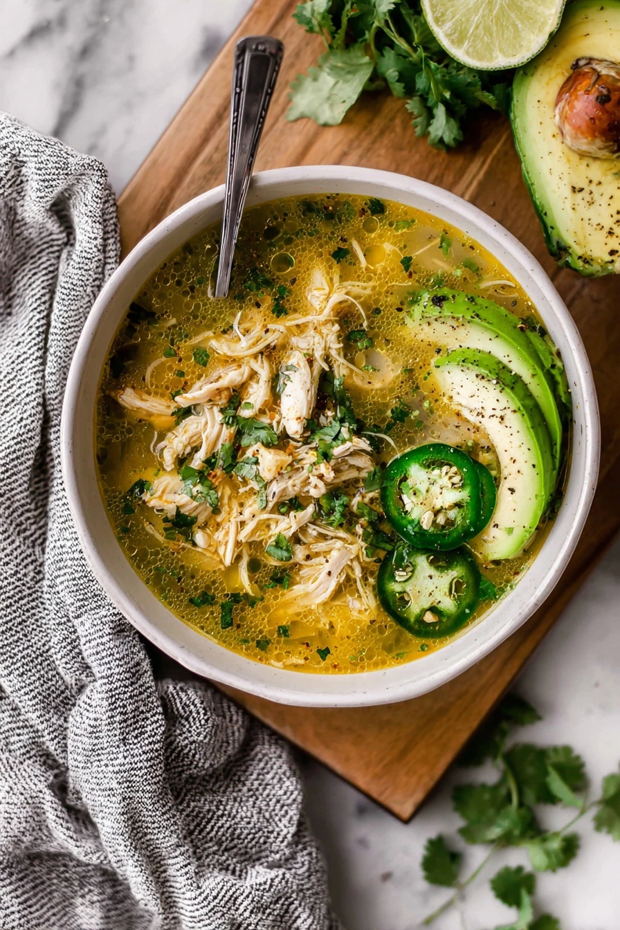A white bowl sits on a wooden board with a blue and white striped cloth nearby, filled with a creamy soup that has shredded light brown chicken pieces mixed with small green herbs and orange bits. On top, there are three green jalapeño rings, a half lime with visible texture, and a sliced avocado fan sprinkled with black pepper and green herbs. A silver spoon leans into the bowl from the right side. Around the board, there are two lime halves and scattered green cilantro leaves on a white marbled surface. Photo taken with an iphone --ar 2:3 --v 7