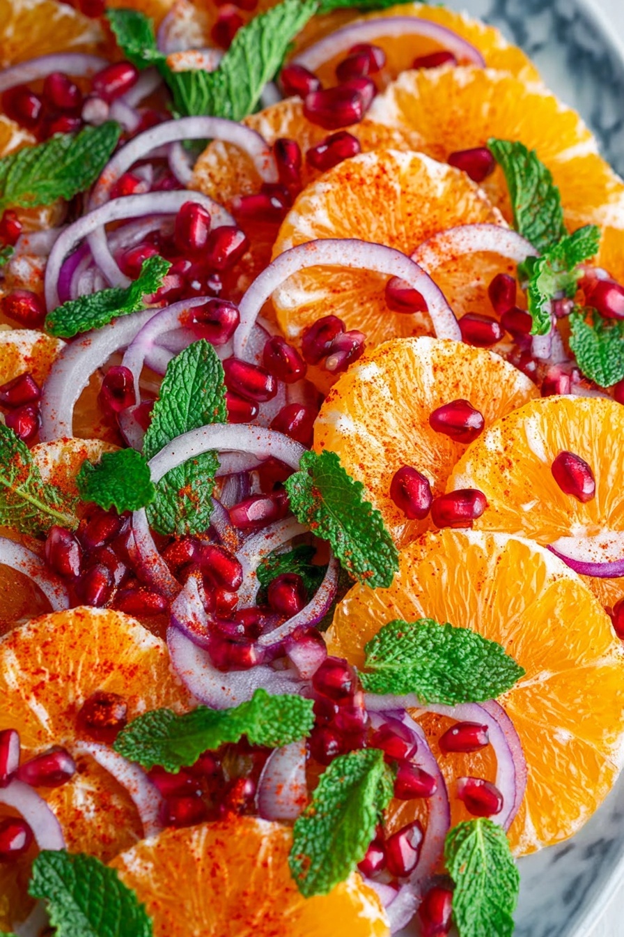 The dish shows thin round layers of orange slices arranged closely inside a white bowl. Over these, there are scattered thin slices of purple onion and bright red pomegranate seeds, adding pops of color. Fresh green mint leaves are spread evenly on top, giving a leafy texture. A light sprinkle of reddish spices dusts the oranges and onions, creating a textured look. The bowl sits on a white marbled surface with some light fabric visible on the side. The photo taken with an iphone --ar 2:3 --v 7