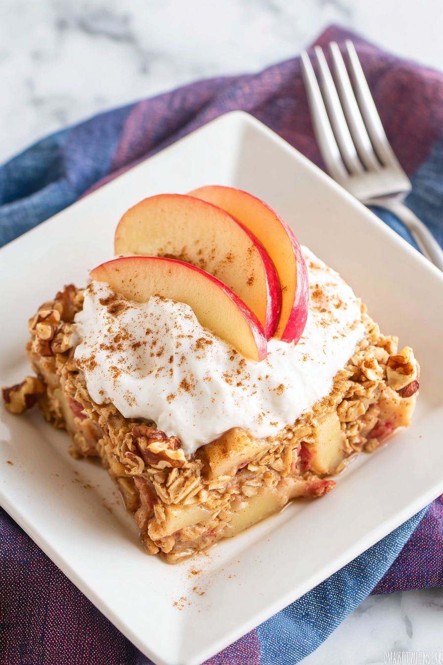 The dish shows a single square piece of apple oatmeal bake on a white square plate. The base layer is a chunky mix of cooked oats, chunks of red-skinned apple, and walnuts with a slightly golden brown color and a soft texture. On top, there is a thick dollop of white whipped cream sprinkled lightly with cinnamon powder, and three thin slices of red apple fanned neatly on one side of the cream. The plate rests on a white marbled surface with a silver fork and folded blue and purple napkins in the background. Photo taken with an iphone --ar 2:3 --v 7