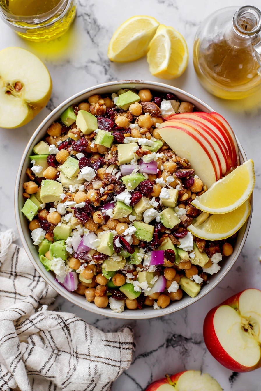 A white bowl full of a colorful salad sits on a white marbled surface. The salad has three main layers: the bottom layer is made of round, light beige chickpeas; the middle layer has chunky pieces of red and green apple and chunks of green avocado mixed evenly. The top layer is scattered with small purple onion cubes, dark red dried cranberries, white crumbled cheese, and little pieces of brown pecans. On the left side of the bowl, there are three yellow lemon wedges placed above the salad. Around the bowl, there is a striped cloth napkin, half a lemon, half a red apple, a wooden board with two green avocado slices, a small glass bottle with light yellow olive oil, and a few pecans scattered. The photo taken with an iphone --ar 2:3 --v 7