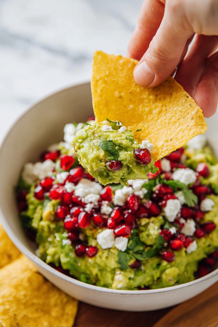 A bowl of guacamole sits in the center of a round wooden board on a white marbled surface, filled with a smooth green avocado base. On top, there are red pomegranate seeds scattered evenly, white crumbled cheese pieces, and fresh green cilantro leaves adding color contrast. A yellow tortilla chip is partially dipped into the guacamole, and several more chips surround the bowl on the board. Some fresh cilantro leaves are placed on the edge of the board. Nearby is a blue cloth napkin. photo taken with an iphone --ar 2:3 --v 7