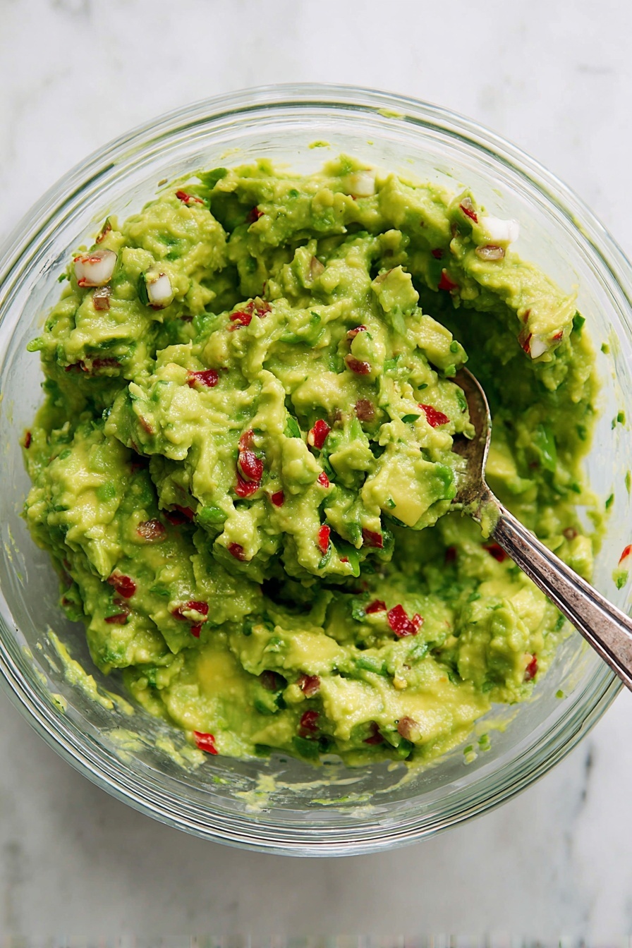 A clear glass bowl filled with a chunky green guacamole that shows bits of red chili peppers, white onion pieces, and finely chopped green herbs mixed throughout. A silver spoon is partially dipped into the guacamole on the right side of the bowl, resting inside it. The bowl sits on a white marbled surface. The guacamole looks fresh with a mix of smooth and textured areas, showing pockets of mashed avocado blended with solid small pieces. Photo taken with an iphone --ar 2:3 --v 7