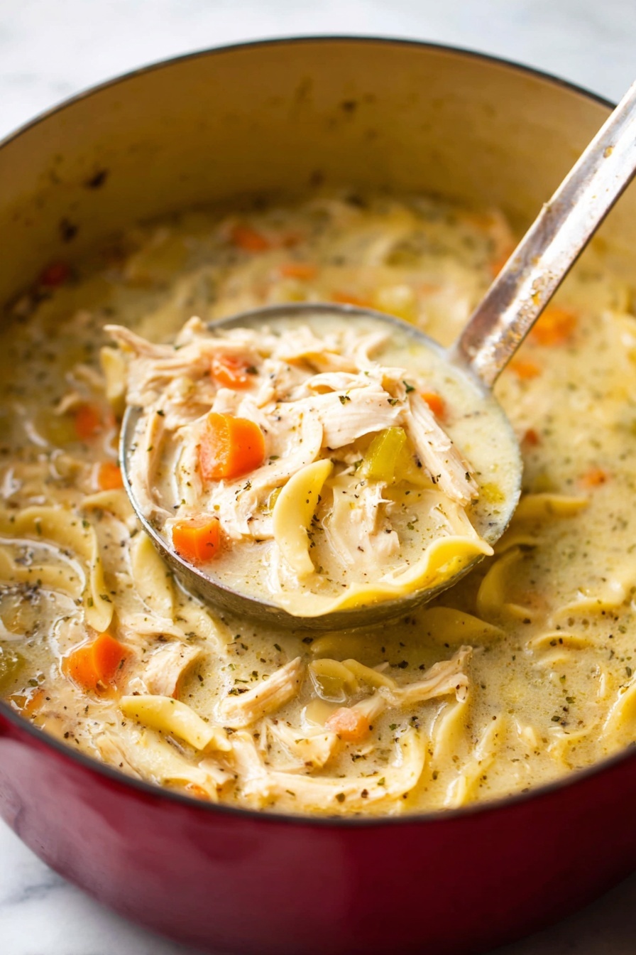 The image shows a close-up of a thick creamy soup in a large red cooking pot with a metal ladle lifting a portion. The soup has a pale beige creamy base full of shredded pieces of light brown chicken, soft yellow noodles, and thin orange carrot slices mixed throughout. The soup surface is speckled with small black seasoning bits, giving it a textured look. The pot sits on a white marbled surface. photo taken with an iphone --ar 2:3 --v 7