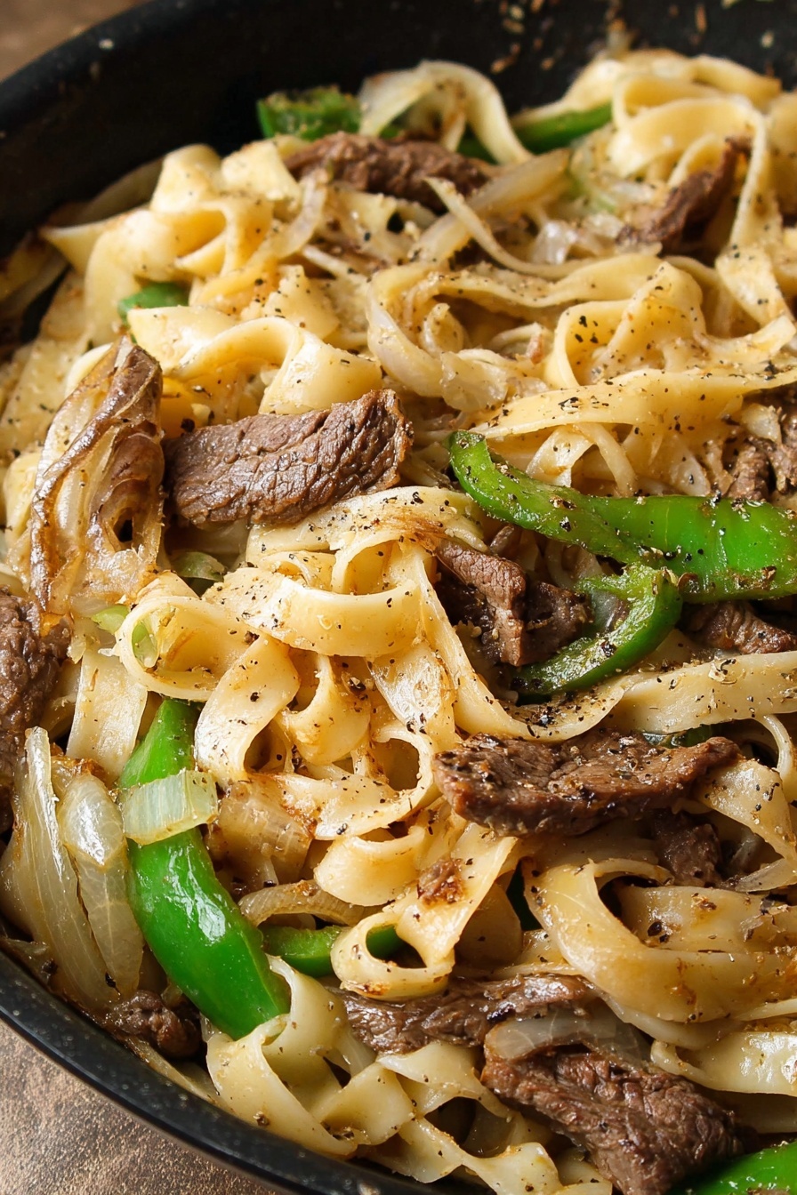 This image shows a black pan filled with a pasta dish that has three clear layers. The bottom layer is wide, light beige pasta ribbons that are soft and slightly curled. The middle layer consists of sautéed green bell pepper slices and translucent, golden brown onion slices, scattered evenly among the pasta. The top layer has small pieces of browned meat sprinkled with black pepper, which rest on the vegetables and pasta. The pan sits on a white marbled surface with a beige cloth partially visible under it. photo taken with an iphone --ar 2:3 --v 7