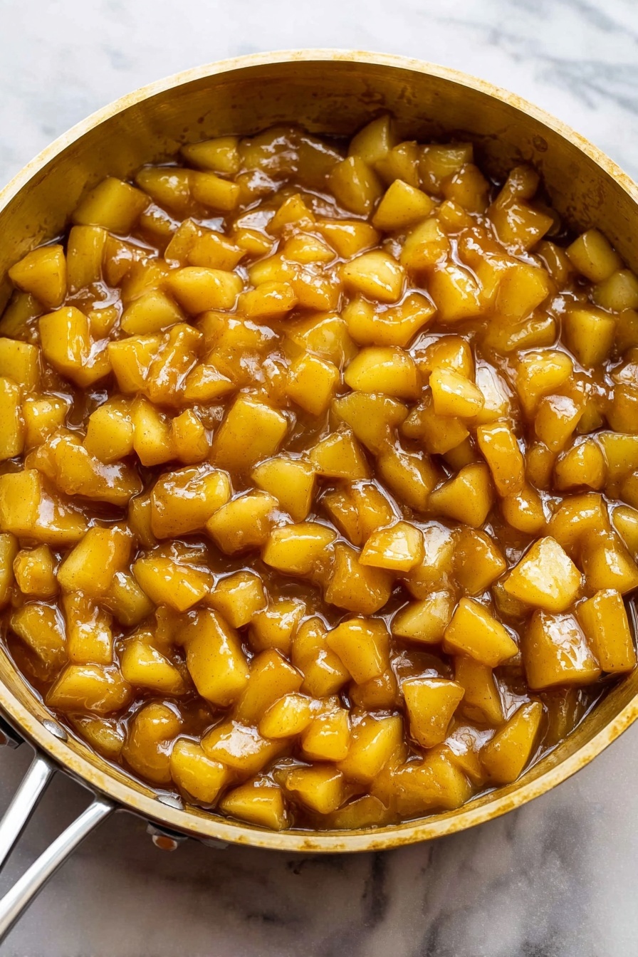 The image shows a close-up of a single layer of small, cubed pieces of cooked apple in a light brown sauce inside a gold-colored pan. The apple pieces are coated evenly with the shiny sauce that has a smooth, slightly thick texture. The cubes of apple vary slightly in size and show a soft, tender appearance with some edges rounded from cooking. The pan is golden metal with visible rivets on the side, and the background surface is changed to a white marbled texture. photo taken with an iphone --ar 2:3 --v 7
