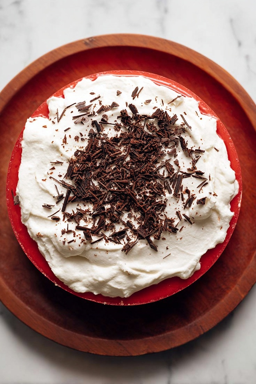 The dessert has three distinct layers placed on a rustic wooden stand with a white marbled background: the bottom layer is dark brown and crumbly, the middle layer is smooth and vibrant red, and the top layer is thick white whipped cream with small dark chocolate shavings sprinkled over it. The red layer looks shiny and firm, while the whipped cream appears soft and fluffy, creating a nice contrast with the chocolate bits on top. Photo taken with an iphone --ar 2:3 --v 7