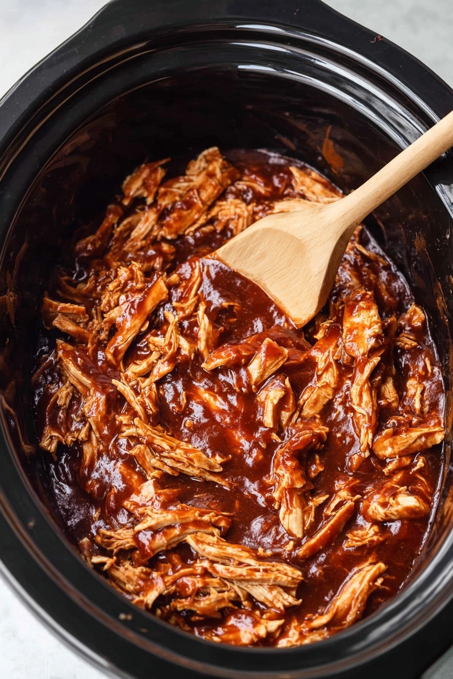 This image shows a close-up of shredded chicken covered in rich, dark reddish-brown barbecue sauce inside a black slow cooker. The chicken pieces are moist and tender, layered unevenly with visible strands and chunks all mixed in the thick, glossy sauce. A wooden spoon with light grain texture rests on the right side, partially submerged in the saucy chicken. The background is softly blurred showing the dark interior of the slow cooker. photo taken with an iphone --ar 2:3 --v 7