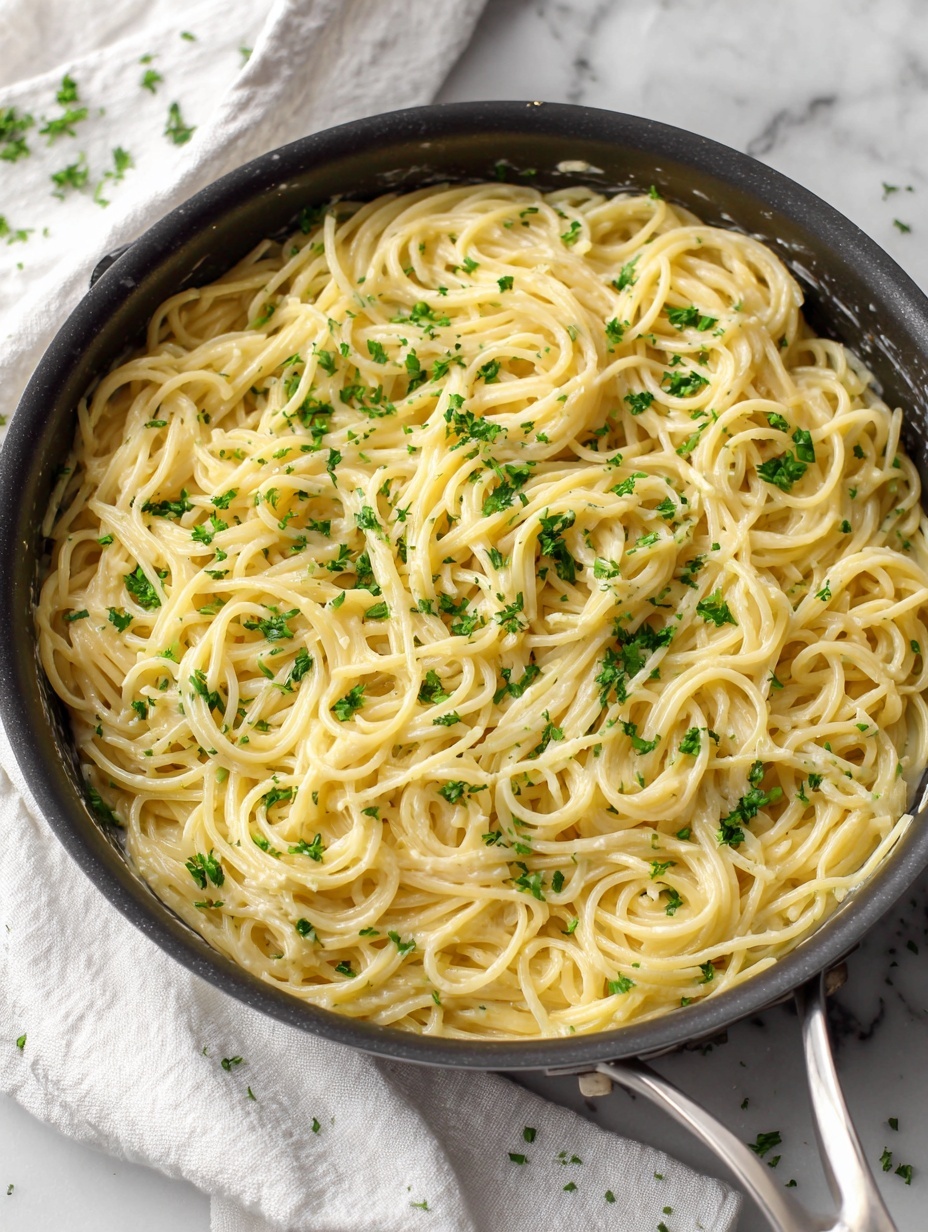 A close-up top view of a large silver pan filled with creamy spaghetti pasta, showing long, smooth, yellow noodles mixed with small green herb pieces evenly spread throughout. Two forks are placed inside, one twirling a portion of spaghetti while the other lifts some noodles, creating soft curves and strands. The pan sits on a white marbled surface with a white cloth partially visible. The sauce coating the pasta is light in color and smooth, giving the noodles a slightly shiny look. photo taken with an iphone --ar 2:3 --v 7