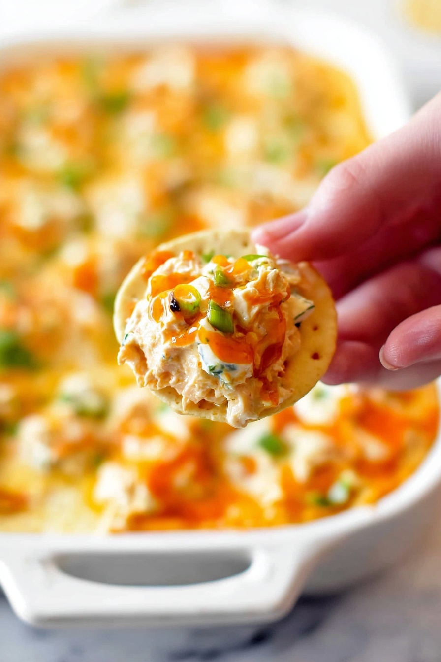 The dish is shown in a white rectangular baking dish filled with three visible layers: the bottom layer is a soft orange cheesy base, topped with splashes of bright red sauce scattered unevenly across the surface. On top of this, there are small crumbles of white and blue cheese, adding texture and creaminess, and the topmost layer is bright green sliced scallions scattered generously all over. A woman's hand is dipping a yellow round chip on the left side into the cheesy, saucy mix. The background is a white marbled texture. Photo taken with an iphone --ar 2:3 --v 7