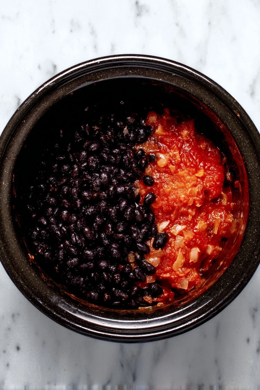 A black slow cooker is filled halfway with two main layers: the bottom layer is a thick, chunky red sauce with small bits of vegetables and seeds, while the top layer is dark, shiny black beans that cover the entire surface above the sauce. The cooker is placed on a white marbled surface. photo taken with an iphone --ar 2:3 --v 7