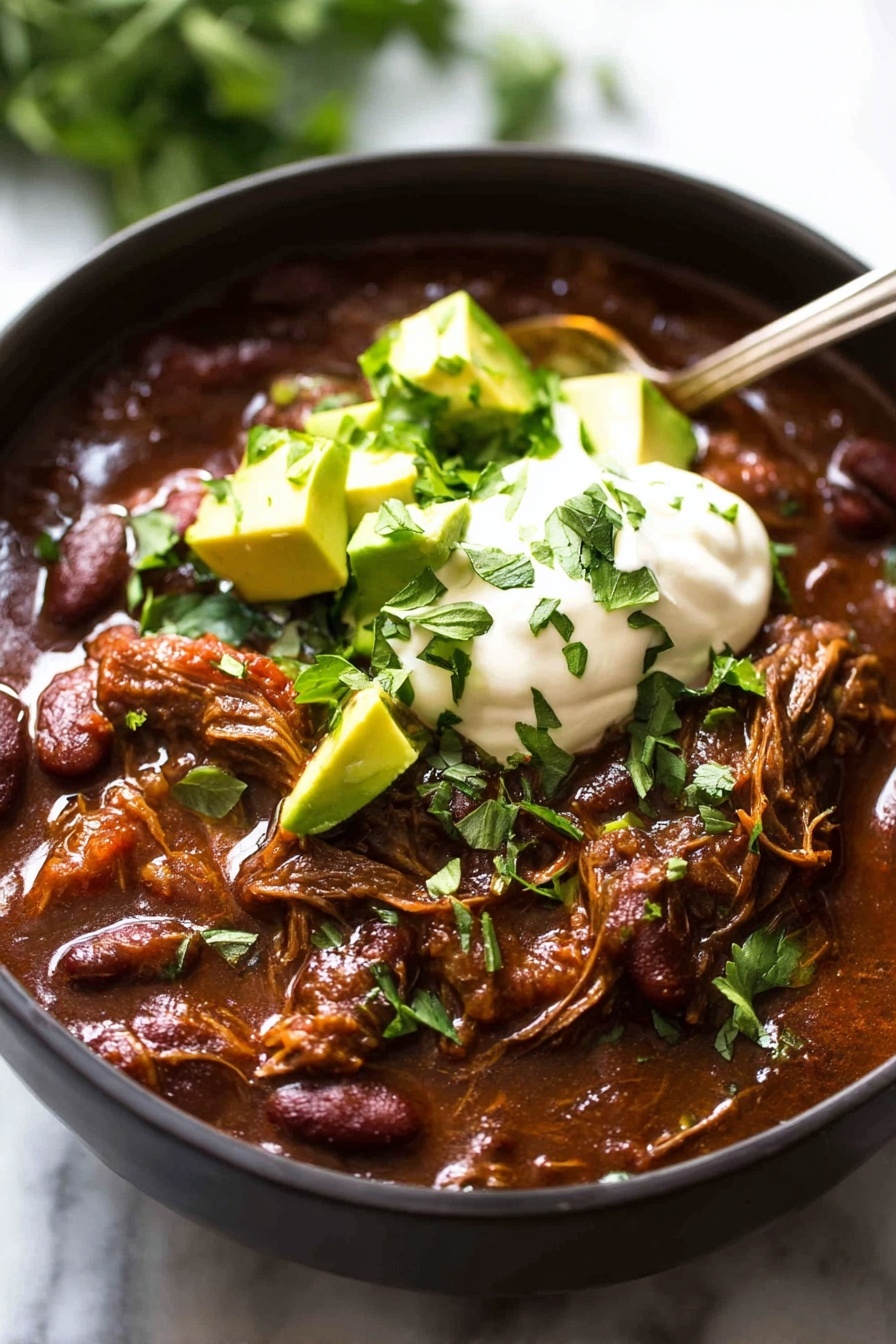 A close-up of a black bowl filled with thick, dark red chili that has visible beans and shredded meat mixed in. On top, there is a dollop of white sour cream, surrounded by small green chunks of avocado and sprinkled with chopped fresh green herbs. A silver spoon rests inside the bowl on the right side. The bowl is placed on a white marbled surface with blurred green leaves in the background. photo taken with an iphone --ar 2:3 --v 7